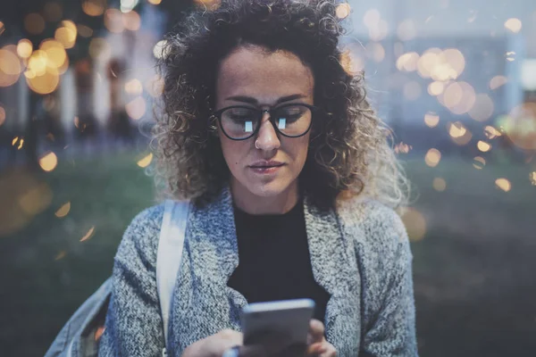 Mujer hipster con mochila y gafas buscando información en la red móvil por teléfono inteligente, durante el paseo por la ciudad europea en la noche. Efectos de bokeh y bengalas sobre fondo borroso . — Foto de Stock