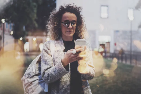 Femme hipster portant un sac à dos et des lunettes de recherche d'informations dans le réseau mobile par téléphone intelligent, lors d'une promenade dans la ville européenne la nuit. Bokeh et effets de fusées éclairantes sur fond flou . — Photo