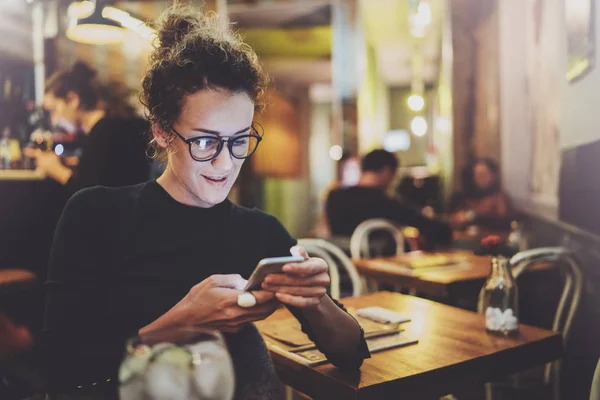 Encantadora mujer con una hermosa sonrisa leyendo un mensaje de texto en el teléfono móvil durante el descanso en la cafetería. Efecto bokeh y bengalas sobre fondo borroso . —  Fotos de Stock