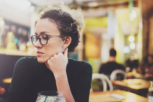 Чарівна модна жінка з окулярами в чорному светрі сидить за столом у кафе вночі. Боке і спалахи впливають на розмитий фон . — стокове фото