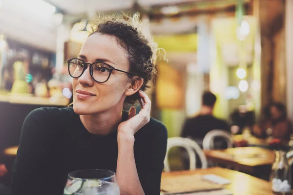 La donna alla moda incantevole con occhiali di occhi in un maglione nero si siede a un tavolo in un caffè di notte. Effetto bokeh e bagliori su sfondo sfocato . — Foto Stock