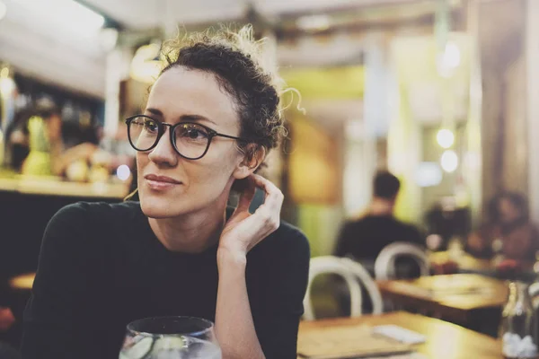 La donna alla moda incantevole con occhiali di occhi in un maglione nero si siede a un tavolo in un caffè di notte. Effetto bokeh e bagliori su sfondo sfocato . — Foto Stock