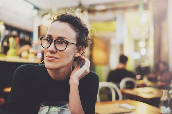 La donna alla moda incantevole con occhiali di occhi in un maglione nero si siede a un tavolo in un caffè di notte. Effetto bokeh e bagliori su sfondo sfocato . — Foto Stock