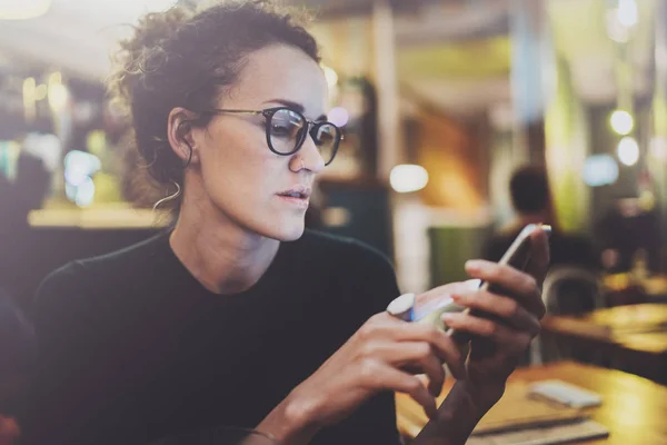 Charmante vrouw bril lezen van e-mailbericht op mobiele telefoon tijdens rust in de coffeeshop. Bokeh en fakkels effect op onscherpe achtergrond. — Stockfoto
