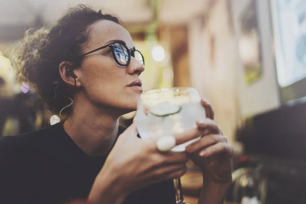 Charming fashionable woman with eyes glasses in a black sweater sits at a table in a cafe at night. Bokeh and flares effect on blurred background. — Stock Photo, Image