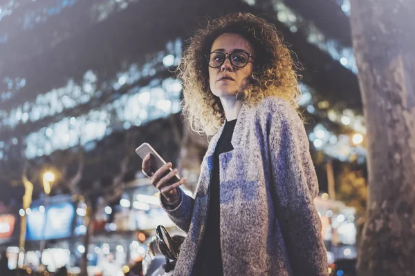 Hipster piękna dziewczyna w okularach trzymając się za ręce inteligentny telefon, podczas nocy spaceru w powołaniu Europejskiego. Efekty bokeh i flary na niewyraźne tło. — Zdjęcie stockowe