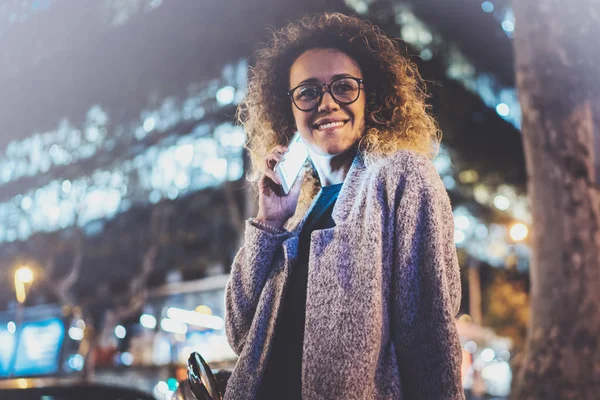 Smiling pretty woman making conversation call with her friends via cell telephone while standing at night on the street. Bokeh and flares effect on blurred background. — Stock Photo, Image