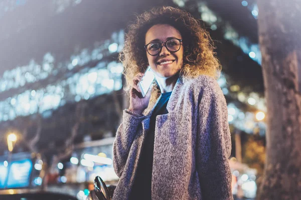 Smiling pretty woman making conversation call with her friends via cell telephone while standing at night on the street. Bokeh and flares effect on blurred background.