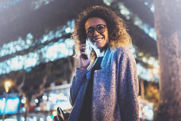 Mosolygó csinos nő így beszélgetés hív keresztül a sejt telefon, éjjel az utcán állva barátaival. Bokeh, fáklya hatása elmosódott háttér. — Stock Fotó