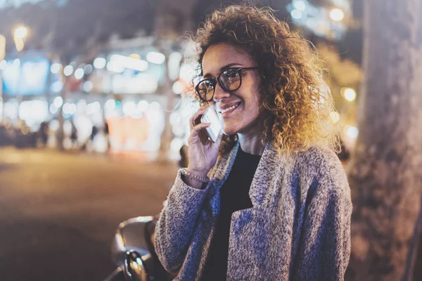 Smiling pretty woman making conversation call with her friends via cell telephone while standing at night on the street. Bokeh and flares effect on blurred background. — Stock Photo, Image