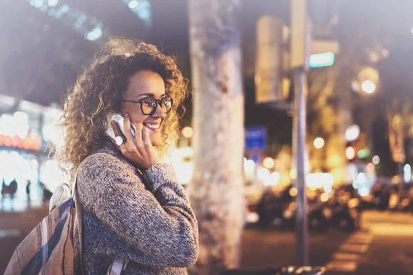 Jolie femme souriante qui passe un appel de conversation avec ses amis par téléphone portable pendant qu'elle se tient debout la nuit dans la rue. Effet Bokeh et fusées éclairantes sur fond flou . — Photo