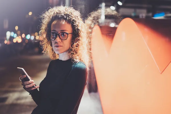 Belle fille hipster portant des lunettes tenant des mains de téléphone intelligent, pendant la marche de nuit dans la vocation européenne. Bokeh et effets de fusées éclairantes sur fond flou . — Photo