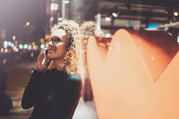 Smiling pretty woman making conversation call with her friends via cell telephone while standing at night on the street. Bokeh and flares effect on blurred background. — Stock Photo, Image
