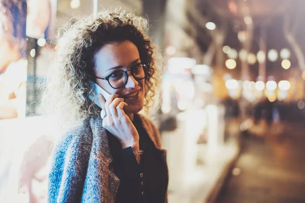 Jolie femme souriante qui passe un appel de conversation avec ses amis par téléphone portable pendant qu'elle se tient debout la nuit dans la rue. Effet Bokeh et fusées éclairantes sur fond flou . — Photo