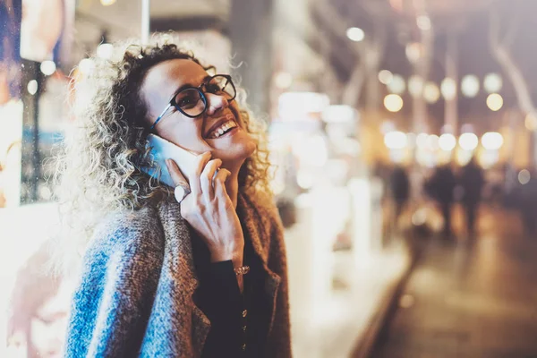 Mujer bonita sonriente haciendo llamadas de conversación con sus amigos a través del teléfono celular mientras está de pie por la noche en la calle. Efecto bokeh y bengalas sobre fondo borroso . — Foto de Stock