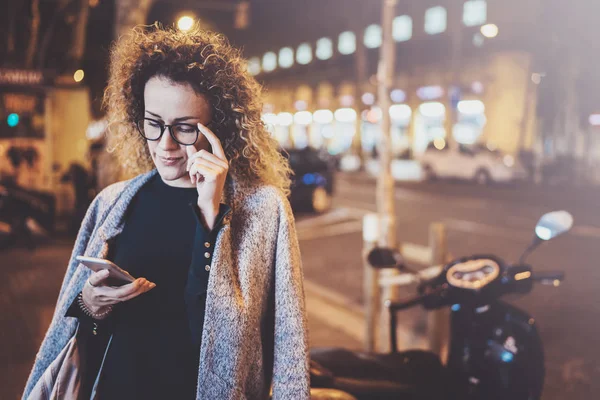 Hermosa chica hipster con gafas sosteniendo las manos del teléfono inteligente, durante la noche caminando en vocación europea. Efectos de bokeh y bengalas sobre fondo borroso . —  Fotos de Stock