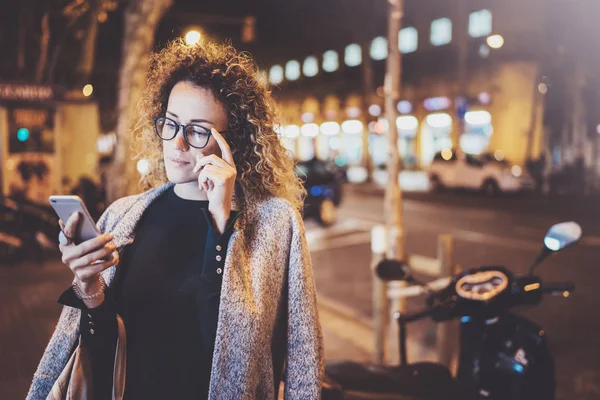 Hermosa chica hipster con gafas sosteniendo las manos del teléfono inteligente, durante la noche caminando en vocación europea. Efectos de bokeh y bengalas sobre fondo borroso . — Foto de Stock