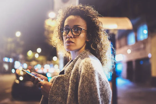 Menina bonita hipster usando óculos segurando as mãos de telefone inteligente, durante a noite andando em vocação europeia. Bokeh e flares efeitos sobre fundo desfocado . — Fotografia de Stock