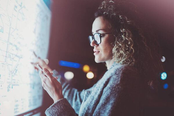 Charming fashionable woman tourist with eyes glasses looking on smartphone while checking way on map of the city on electronic bulletin board. Bokeh and flares effect on blurred background.