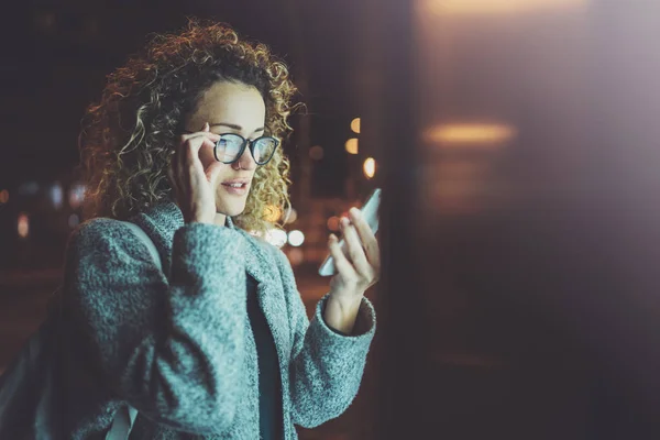 Vrouw in bril houden handen smartphone in de sfeervolle stad nacht. Vrouwelijke handen met behulp van de mobiele telefoon. Close-up op onscherpe achtergrond. Fakkels, bokeh effecten. — Stockfoto