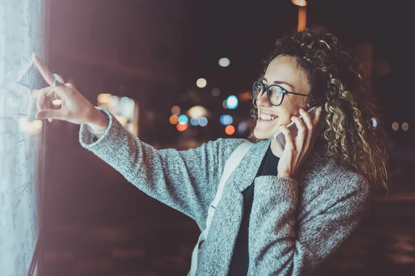 Feliz turista de mulher na moda com óculos de olhos que falam no telefone celular enquanto olha para o mapa da cidade no quadro de avisos eletrônico. Bokeh e flares efeito sobre fundo desfocado . — Fotografia de Stock