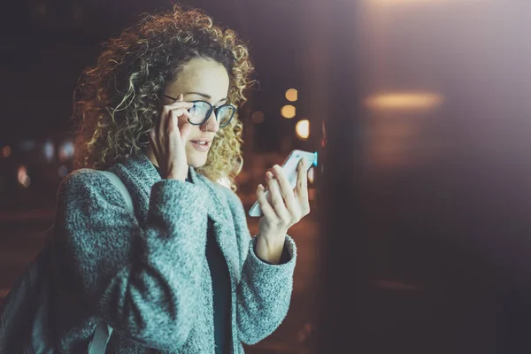 Mulher em óculos de olho de mãos dadas smartphone na cidade atmosférica noturna.Mãos femininas usando telefone móvel.Fechar-se em fundo borrado .Clarissas, efeitos bokeh . — Fotografia de Stock