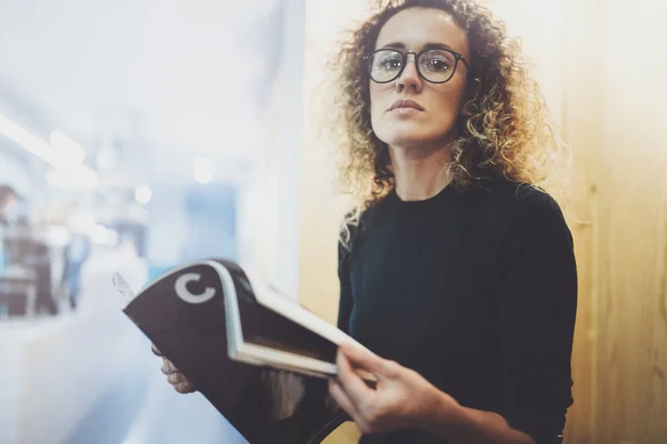 Charming fashionable woman with eyes glasses reading magazine sitting indoor in urban cafe.Casual portrait of pretty girl. Blurred background. — Stock Photo, Image