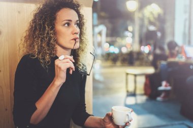 Gece ve kitap okuma bir kafede bir masada bir siyah kazak gözler gözlük ile büyüleyici moda kadın oturur. Arka plan bulanık.