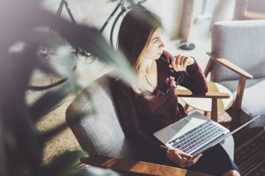 Rahat koltuk oturma ve dizüstü bilgisayar kullanan genç güzel sarışın bir kadın. Studio coworking, işlem çalışma. Arka plan bulanık panoramik pencereler.