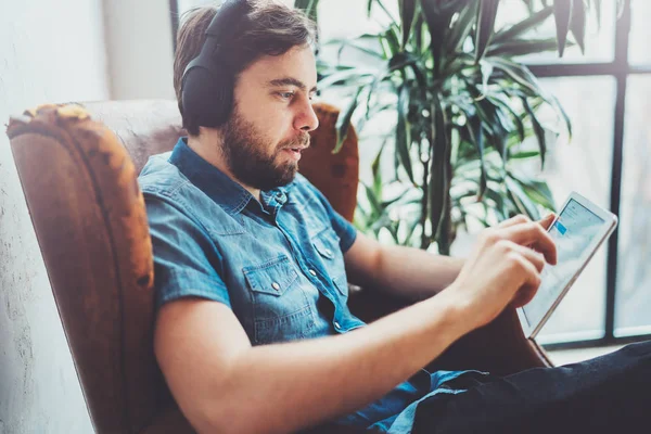 Joven hipster llevar ropa casual escuchando música en los auriculares en la tableta táctil en el hogar moderno place.Horizontal.Blurred fondo . — Foto de Stock