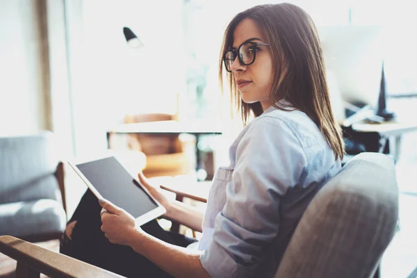 Belle jeune blonde portant des lunettes et utilisant une tablette électronique tactile sur un lieu de travail ensoleillé. Fond flou . — Photo