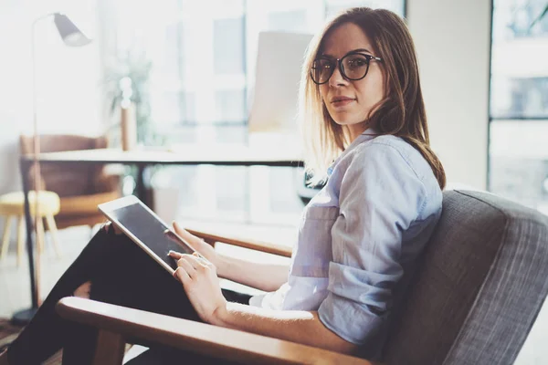 Όμορφος νεαρή ξανθιά φορώντας γυαλιά και χρησιμοποιώντας ηλεκτρονικά touch υπολογιστή tablet στο ηλιόλουστο χώρο εργασίας. Οριζόντια. Θολή φόντο. — Φωτογραφία Αρχείου