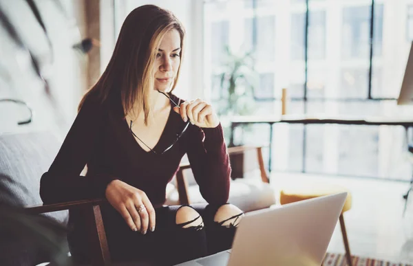 Ung vacker blond kvinna sitter på en bekväm fåtölj och använder bärbar dator. Arbetar process på coworking studio. Panoramafönster på suddig bakgrund. — Stockfoto