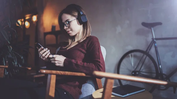 Joven chica hipster atractiva con gafas para los ojos y ropa casual escuchando música en los auriculares en el teléfono inteligente en el lugar moderno . — Foto de Stock