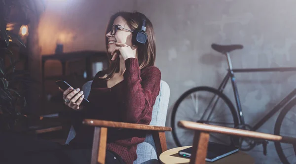 Chica atractiva joven con gafas para los ojos y ropa casual escuchando música en los auriculares en el teléfono inteligente en el lugar moderno . — Foto de Stock