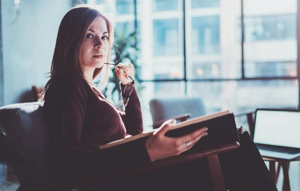 Stilig ung flicka bär glasögon casual kläder håller boken händer. Kvinna blondin sitter i vintage fåtölj moderna loft studio läser bok. Suddig bakgrund. Horisontell. — Stockfoto