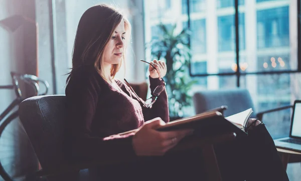 Knap jong meisje dragen casual kleding glazen boek handen te houden. Vrouw blond zit in vintage fauteuil moderne loft studio lezen boek. Onscherpe achtergrond. Horizontale. — Stockfoto