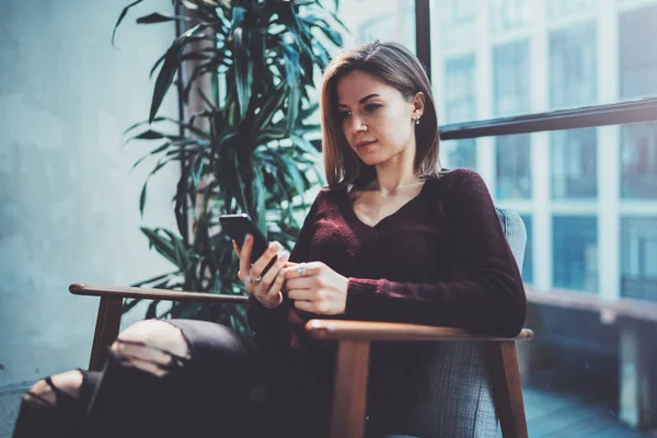 Blonde yound vrouw met moderne mobiele telefoon in handen. Meisje wijzen vingers op lege mobiele aanraakscherm op zonnige naaiatelier plaats. Horizontale, onscherpe achtergrond. — Stockfoto