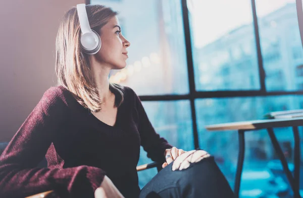 Piękna blondynka na sobie okulary i na co dzień ubrania słuchania muzyki cyfrowej w słuchawkach na smartfonie podczas relaksu w fotel w miejscu nowoczesny. — Zdjęcie stockowe