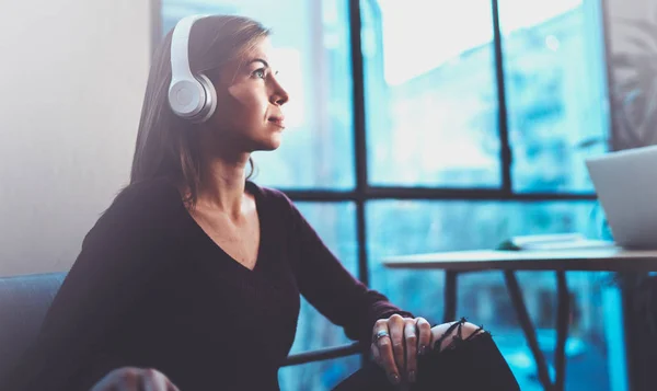 Menina loira bonita usando óculos e roupas casuais ouvindo música digital em fones de ouvido no smartphone enquanto relaxa em poltrona no lugar moderno . — Fotografia de Stock