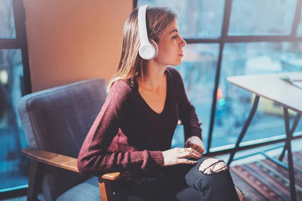 Hermosa chica rubia con gafas para los ojos y ropa casual escuchando música digital en los auriculares en el teléfono inteligente mientras se relaja en el sillón en un lugar moderno . — Foto de Stock