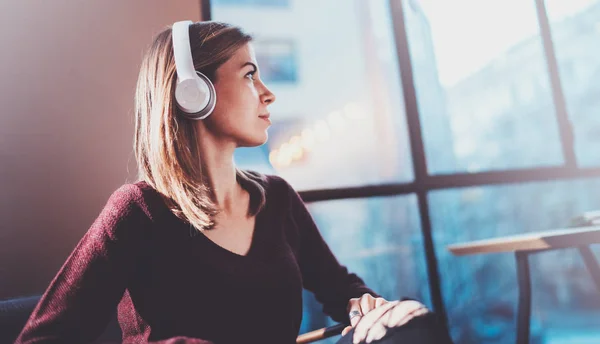 Menina loira bonita usando óculos e roupas casuais ouvindo música digital em fones de ouvido no smartphone enquanto relaxa em poltrona no lugar moderno . — Fotografia de Stock