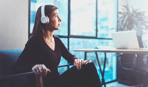 Hermosa chica rubia con gafas para los ojos y ropa casual escuchando música digital en los auriculares en el teléfono inteligente mientras se relaja en el sillón en un lugar moderno . —  Fotos de Stock