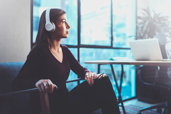 Mooi blond meisje dragen van bril en casual kleding luisteren digitale muziek in de koptelefoon op smartphone terwijl u ontspant in fauteuil op moderne plaats. — Stockfoto