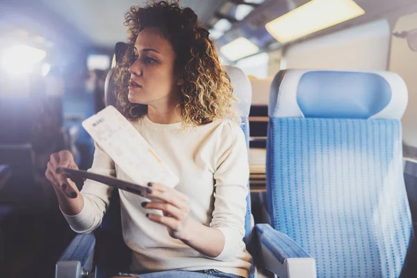 Bénéficiant d'un concept de voyage d'affaires. Jeune belle fille touristique brune voyageant dans le train assis près de la fenêtre à l'aide d'un smartphone, tenant les mains de billets . — Photo