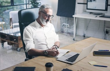 Modern ofis yerinde ahşap masada otururken mobil dizüstü bilgisayar üzerinde çalışan emin yetişkin iş adamı.
