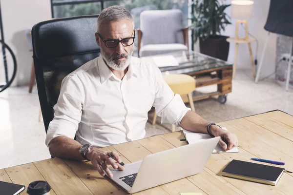 Взрослый бородатый мужчина, работающий за мобильным ноутбуком, сидя за деревянным столом . — стоковое фото