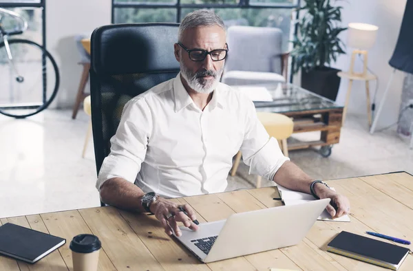 Дорослий бородатий бізнесмен працює на мобільному ноутбуці, сидячи за дерев'яним столом . — стокове фото