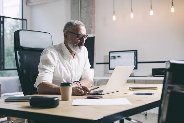 Pengusaha usia pertengahan yang bekerja pada laptop mobile sambil duduk di meja kayu di kantor modern . — Stok Foto