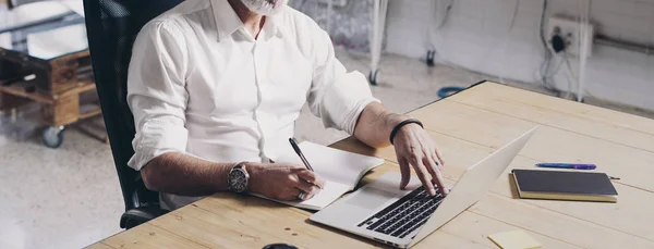 Hombre de negocios adulto positivo usando el ordenador portátil móvil mientras está sentado en la mesa de madera en el moderno lugar de coworking. Amplio. Cultivado . — Foto de Stock